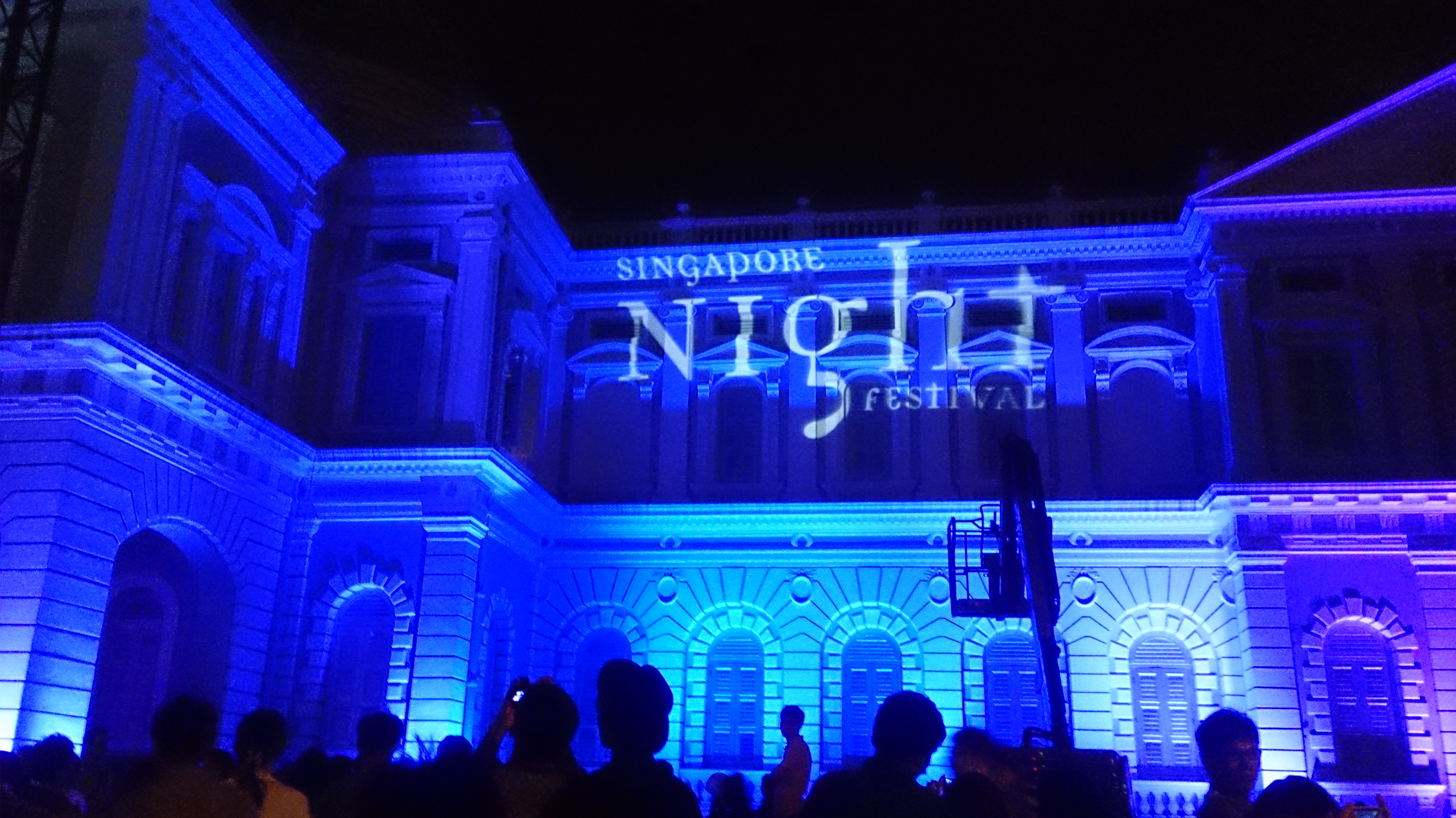 The National Museum of Singapore lit up in deep blue with 'Singapore Night Festival' projected on its façade, with crowds gathered below — the precinct comes alive after dark